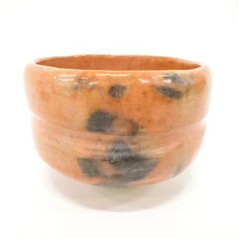 Front view of the Matsukaze Red Raku matcha bowl with persimmon-red glaze, smoky charcoal markings, horizontal ridges, and a pale foot.