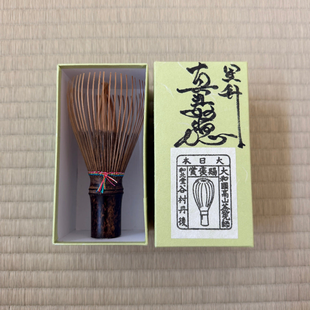 A traditional Japanese bamboo matcha whisk displayed next to its light green presentation box on a beige woven straw mat background.