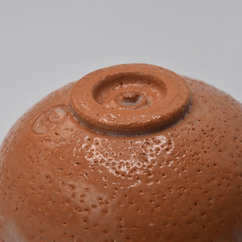 Underside view of the reddish-orange matcha bowl showing its textured surface, raised circular foot, and impressed mark.