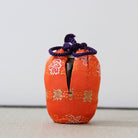 An orange fabric-covered bottle or vase with a drawstring closure, featuring white and gold floral patterns, standing on a light textured surface against a plain light gray background.