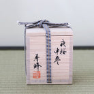 A light natural wood gift box with black Japanese calligraphy and a red seal, tied with a blue-and-white striped ribbon, photographed on a textured green surface against a plain light background.