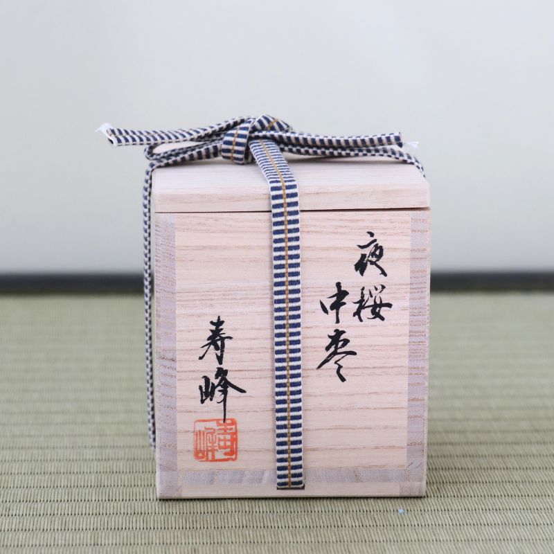 A light natural wood gift box with black Japanese calligraphy and a red seal, tied with a blue-and-white striped ribbon, photographed on a textured green surface against a plain light background.