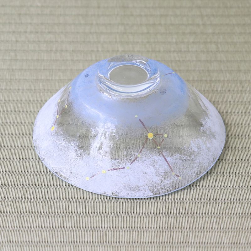 A clear glass bowl with a white and light blue painted design and small yellow celestial dots, photographed from above on a beige woven mat.