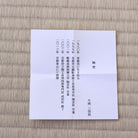 A Japanese condolence card (shitsuregai) with black kanji text on a white paper background, placed flat on a light beige woven straw mat.