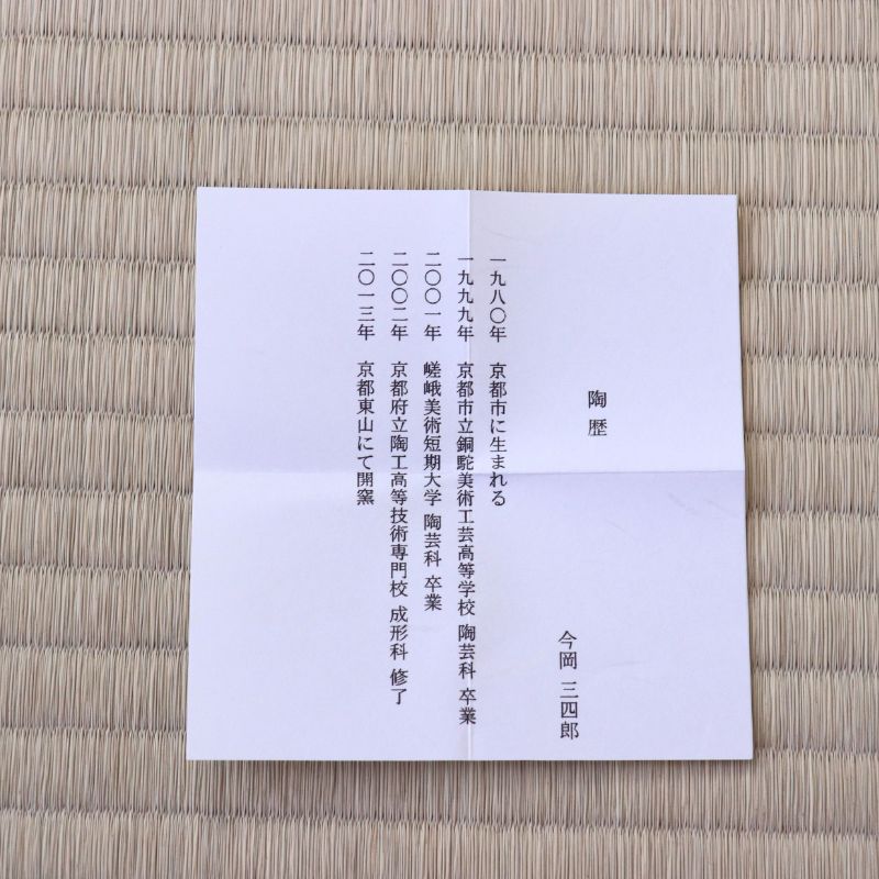 A Japanese condolence card (shitsuregai) with black kanji text on a white paper background, placed flat on a light beige woven straw mat.