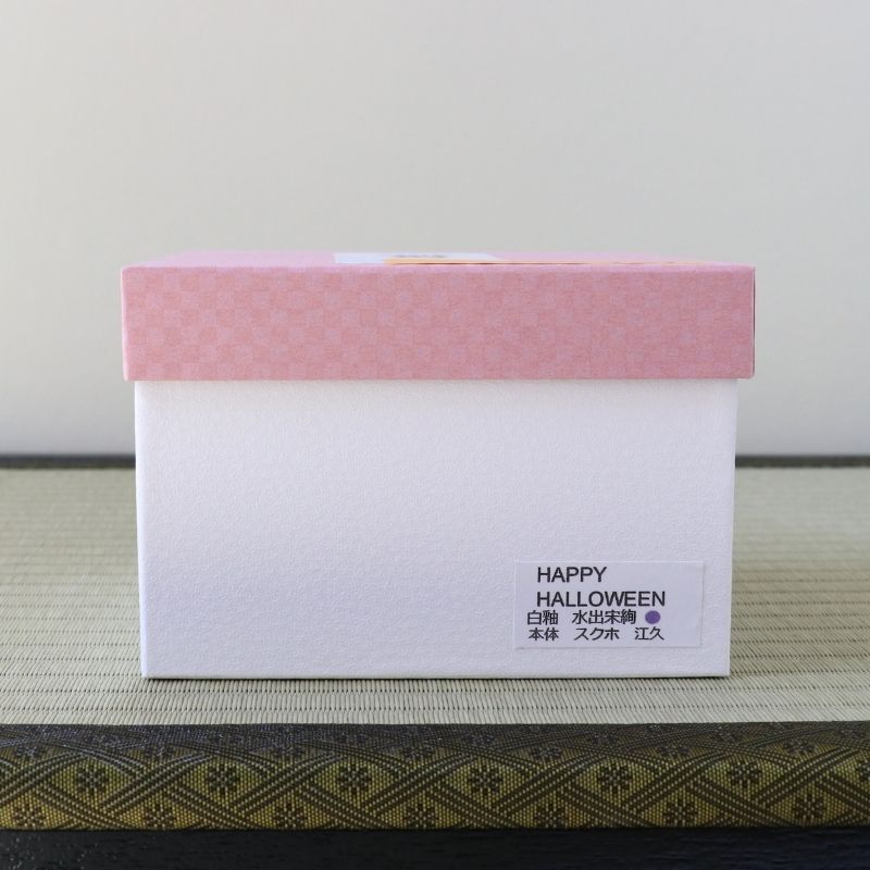 A white rectangular box with a pink patterned lid sits on a textured surface. A small label on the front reads “HAPPY HALLOWEEN” with Japanese text below.