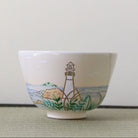 A white ceramic bowl featuring a hand-painted illustration of a lighthouse on a rocky shore with green foliage and blue water, set against a neutral background.