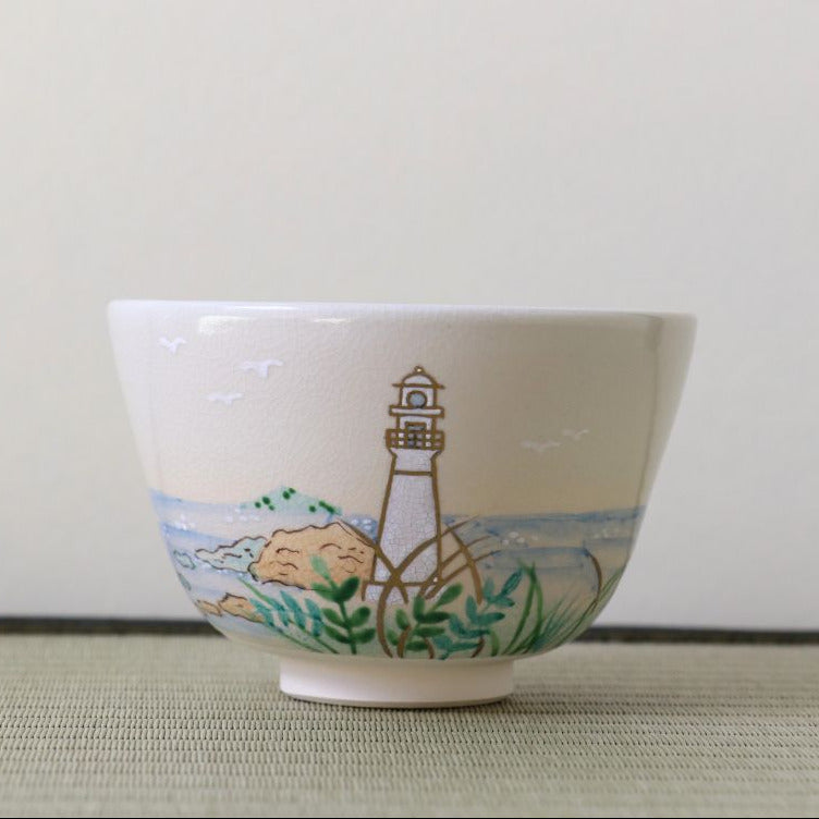 A white ceramic bowl featuring a hand-painted illustration of a lighthouse on a rocky shore with green foliage and blue water, set against a neutral background.