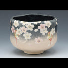 A ceramic tea bowl with a black and beige gradient glaze, decorated with white and pink cherry blossom flowers and small red accents, set against a dark gray background.