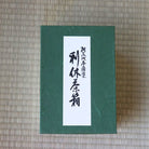 A dark green rectangular box with a white vertical label featuring black Japanese calligraphy, placed on a woven straw mat background.