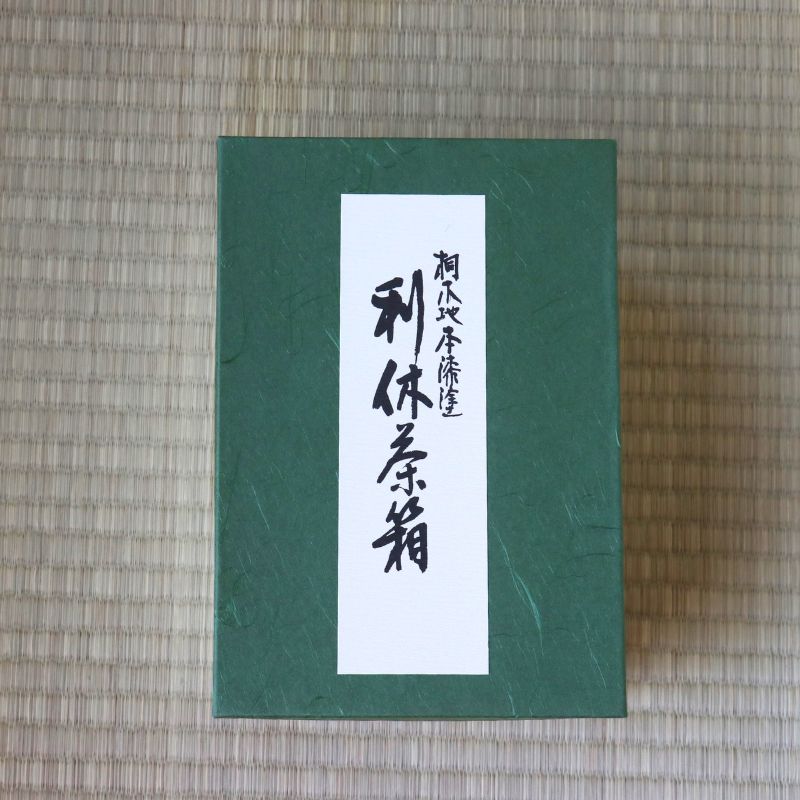 A dark green rectangular box with a white vertical label featuring black Japanese calligraphy, placed on a woven straw mat background.