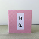 A square pink box with a subtle textured pattern and a white vertical label featuring black Japanese characters, set against a neutral light gray background and a woven mat surface.
