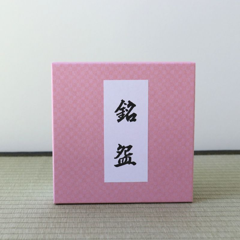 A square pink box with a subtle textured pattern and a white vertical label featuring black Japanese characters, set against a neutral light gray background and a woven mat surface.