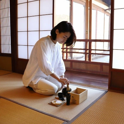 Woman in a traditional white outfit sitting on the floor in a Japanese-style room with tatami mats.