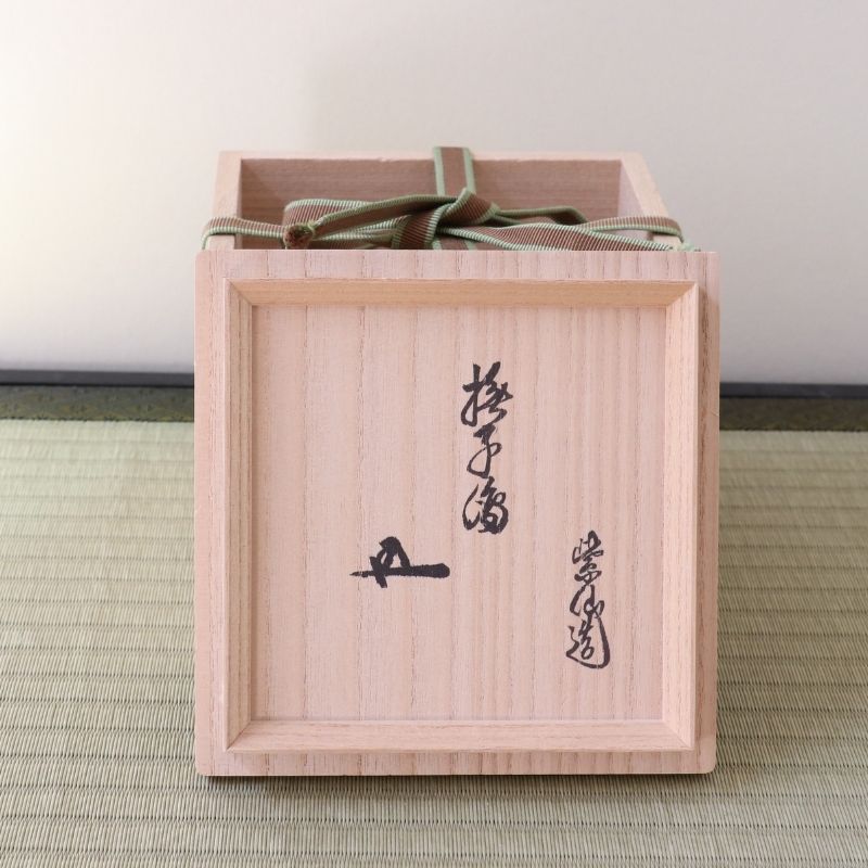 A light natural wood presentation box with black calligraphy markings on the front, tied with a green ribbon, set on a textured green mat against a neutral background.