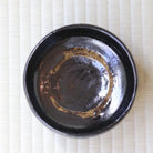 A round black ceramic bowl with a glossy finish and a decorative gold-brown ring near the rim, photographed from directly above on a light beige textured mat.