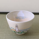 A white ceramic bowl decorated with a hand-painted lighthouse and coastal landscape scene, resting on a woven mat against a neutral background.