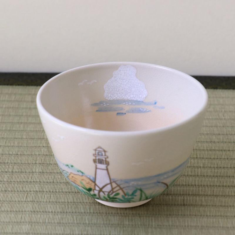 A white ceramic bowl decorated with a hand-painted lighthouse and coastal landscape scene, resting on a woven mat against a neutral background.