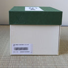 A square product box with a dark green lid and a cream-colored body, placed on a woven mat. A white label with Japanese characters is visible on the lid, and a barcode label is attached to the side.