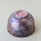 A dark brown ceramic bowl with hand-painted blue floral patterns and gold leaf accents, shown upside down on a light beige woven fabric background.