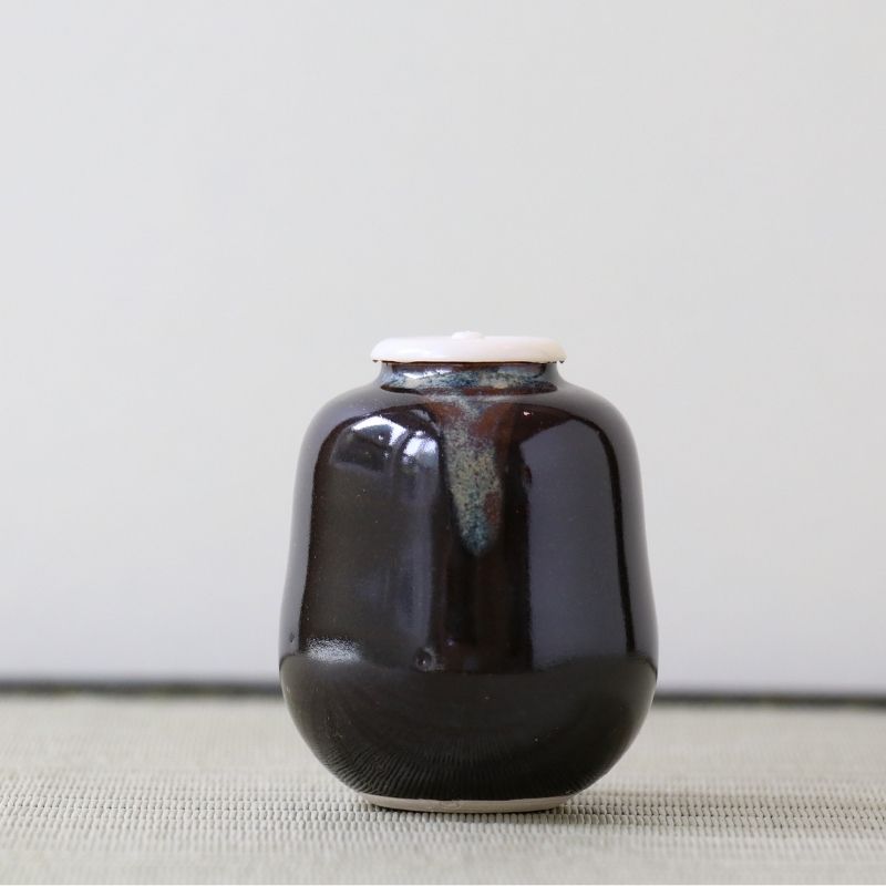A small glossy ceramic jar with a rounded body and a light-colored lid, shown on a neutral textured surface against a plain light background.