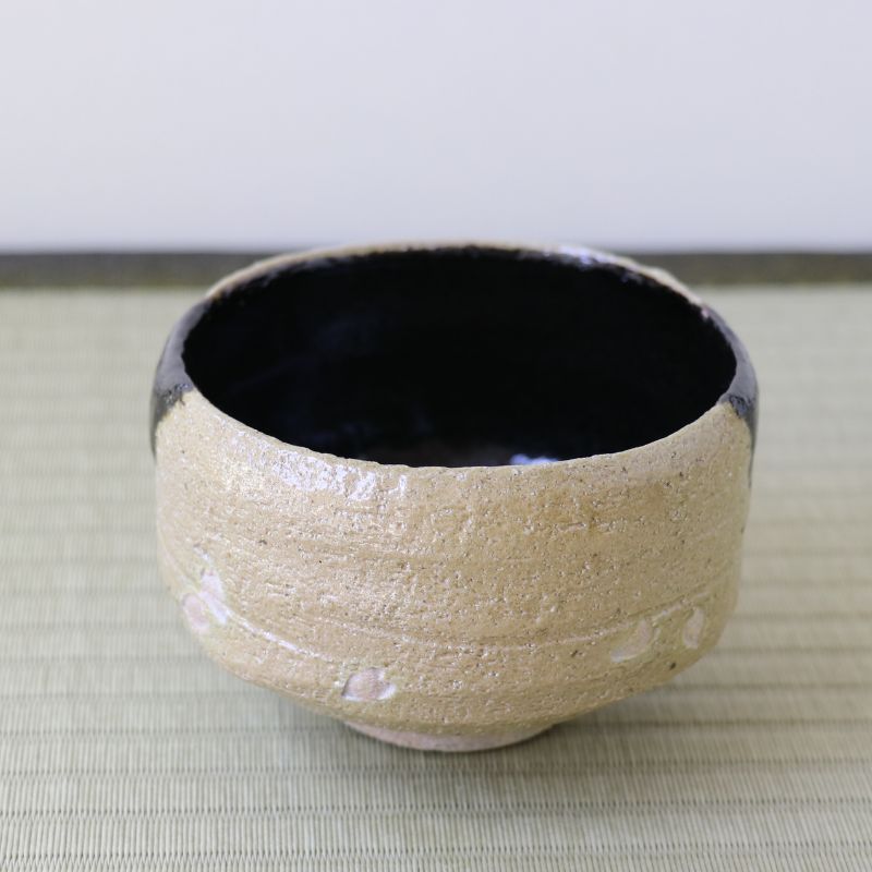 A handmade ceramic tea bowl with a textured beige exterior and a glossy black interior, resting on a light green woven mat.