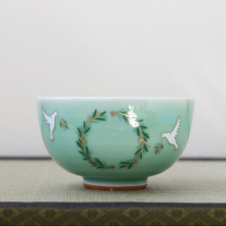 A light green ceramic bowl with hand-painted white doves and a green wreath design with gold accents, resting on a textured surface against a neutral background.