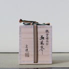 A light natural wood Japanese box with black calligraphy markings and a dark green and beige striped paper cord tied around it, set against a neutral light gray background.
