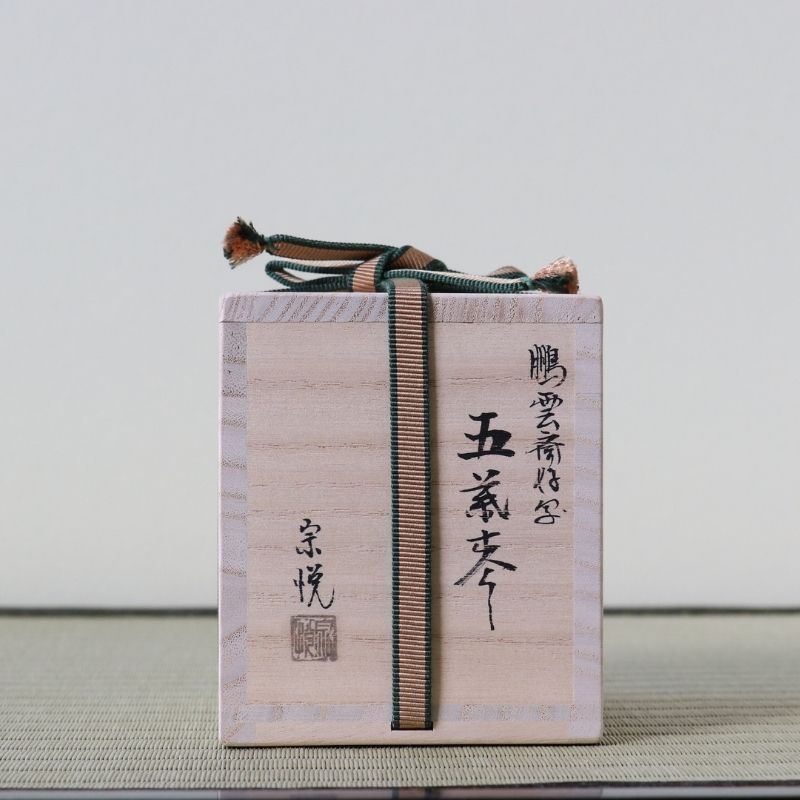 A light natural wood Japanese box with black calligraphy markings and a dark green and beige striped paper cord tied around it, set against a neutral light gray background.