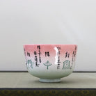 A ceramic tea bowl with a pink and white gradient glaze, decorated with black Japanese calligraphy and small illustrations, resting on a textured beige mat.
