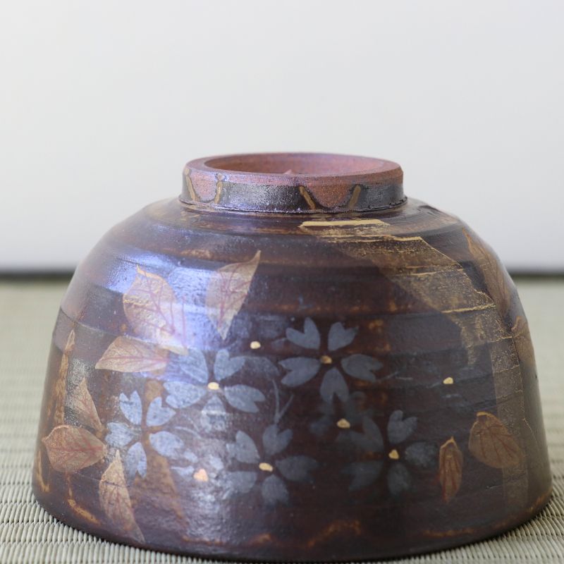 A handmade ceramic bowl with a dark brown and black textured glaze, decorated with hand-painted blue-gray flowers and brown leaves, set on a woven mat against a neutral background.