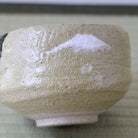 A close-up of a handmade ceramic tea bowl with a textured, speckled glaze in beige and cream tones, featuring a white painted mountain motif on the side.