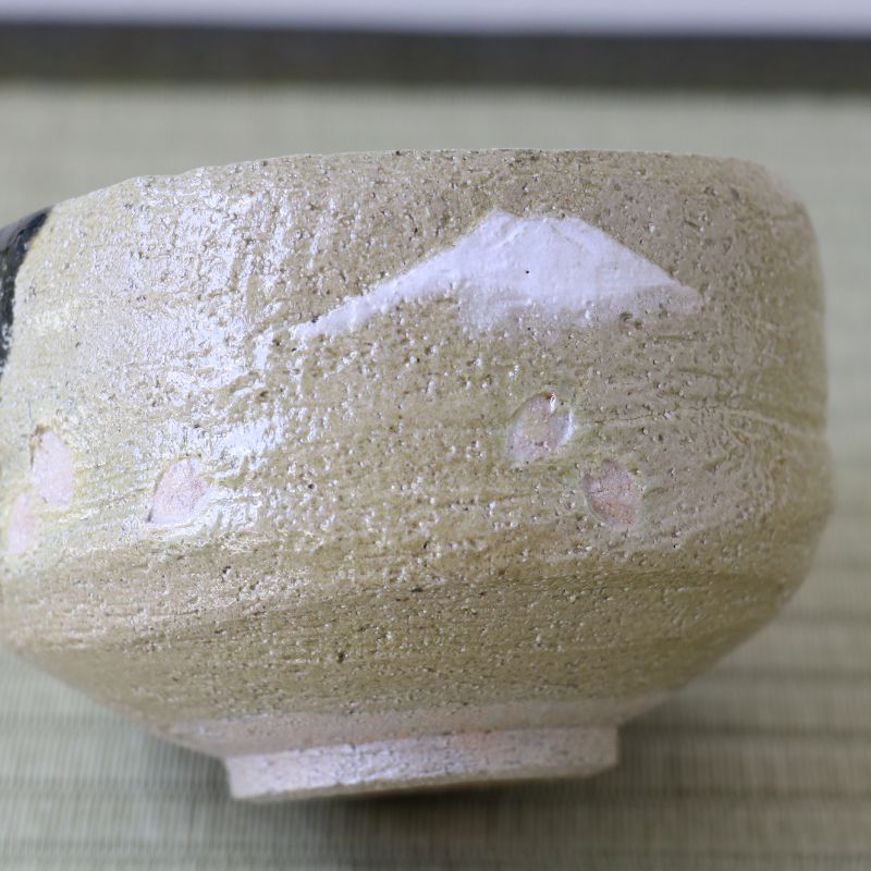 A close-up of a handmade ceramic tea bowl with a textured, speckled glaze in beige and cream tones, featuring a white painted mountain motif on the side.