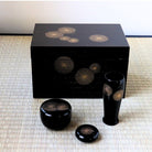 A black lacquerware set with gold sunburst patterns, including a rectangular box, a tall vase, a round lidded container, and a small dish, arranged on a woven mat.