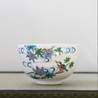 A white ceramic bowl decorated with hand-painted blue and green maple leaves, wavy blue lines, and a small bluebird, set against a neutral light gray background.
