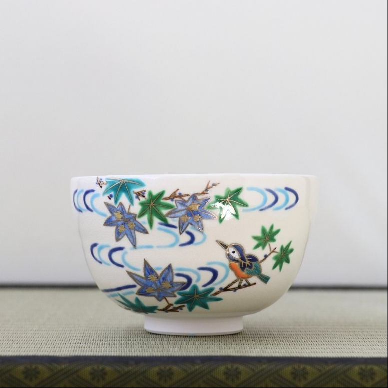 A white ceramic bowl decorated with hand-painted blue and green maple leaves, wavy blue lines, and a small bluebird, set against a neutral light gray background.