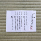 A white rectangular paper document with Japanese text lies on a textured tan woven mat. The paper has black printed text, a large vertical black character on the right, and a red circular stamp near the lower right.