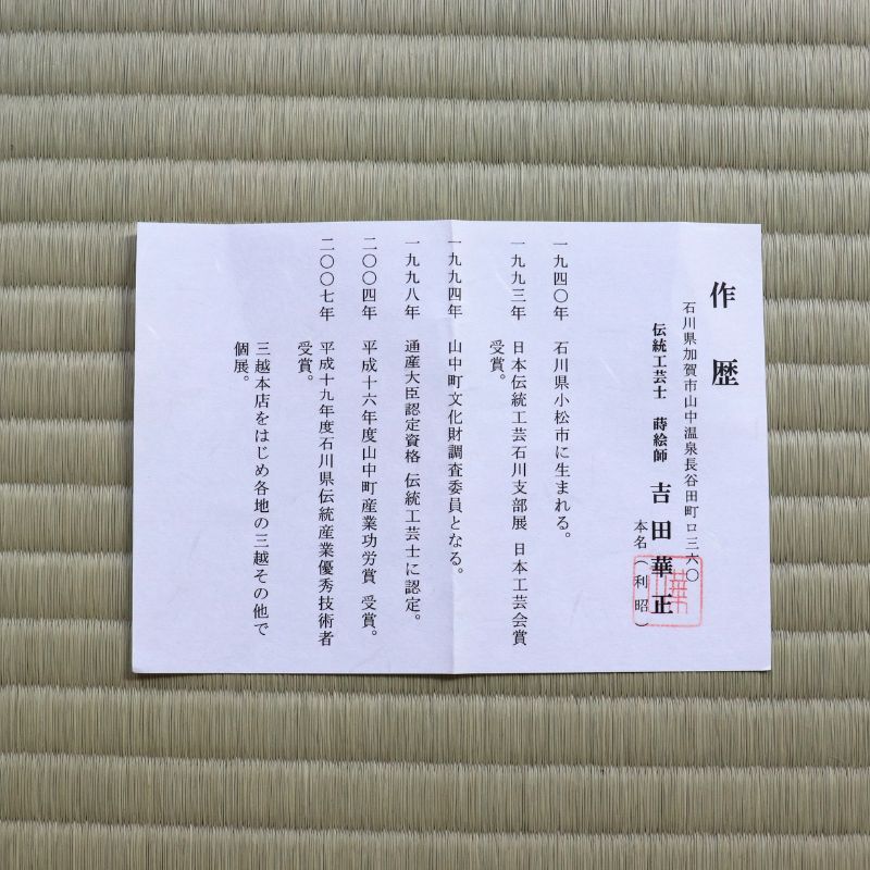 A white rectangular paper document with Japanese text lies on a textured tan woven mat. The paper has black printed text, a large vertical black character on the right, and a red circular stamp near the lower right.