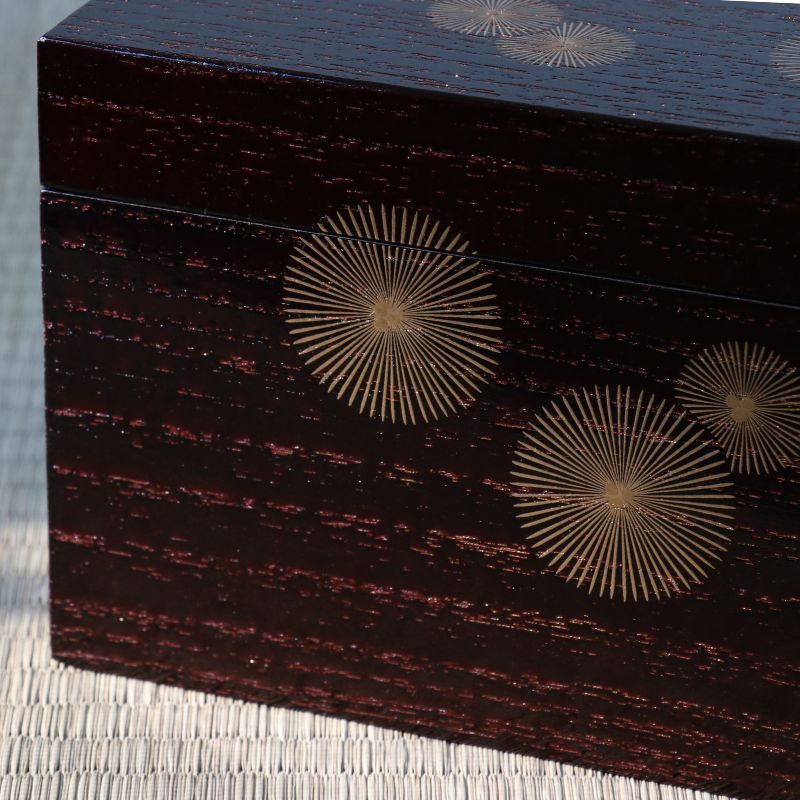 Close-up of a dark reddish-brown lacquered wooden box with gold sunburst inlay patterns on the lid, resting on a woven mat.