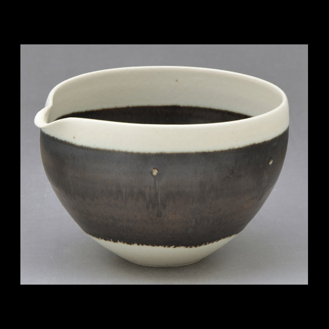 A handmade ceramic bowl with a glossy dark brown glaze and a contrasting matte off-white rim and base, photographed against a neutral gray background.
