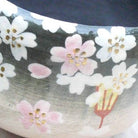 Close-up of a ceramic bowl featuring hand-painted cherry blossoms in white and pink with gold centers on a dark and light gray gradient background.