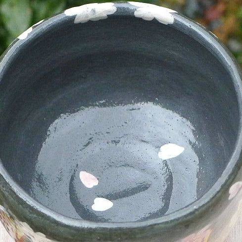 A close-up of a dark glazed ceramic bowl with white and pink floral patterns around the rim, set outdoors on a light-colored surface with blurred green foliage in the background.
