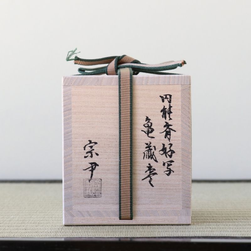 A light natural wood box with black Japanese calligraphy and a striped dark green and brown ribbon tied around it, photographed against a plain light background.