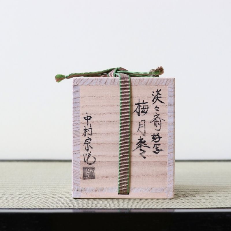 A small light wood gift box with black Japanese calligraphy and a green striped ribbon tied around it, placed on a woven mat against a plain light background.
