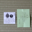 A Japanese tea ceremony tea bag packet (kaiseki-ura) with its white paper label card, displayed on a woven straw mat. The packet is light green with black calligraphy, and the label card features black illustrations and Japanese text.