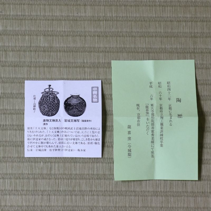 A Japanese tea ceremony tea bag packet (kaiseki-ura) with its white paper label card, displayed on a woven straw mat. The packet is light green with black calligraphy, and the label card features black illustrations and Japanese text.