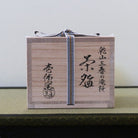 A light natural wood tea box with black Japanese calligraphy and a black-and-white striped ribbon tied around it, placed on a green textured surface against a plain light background.