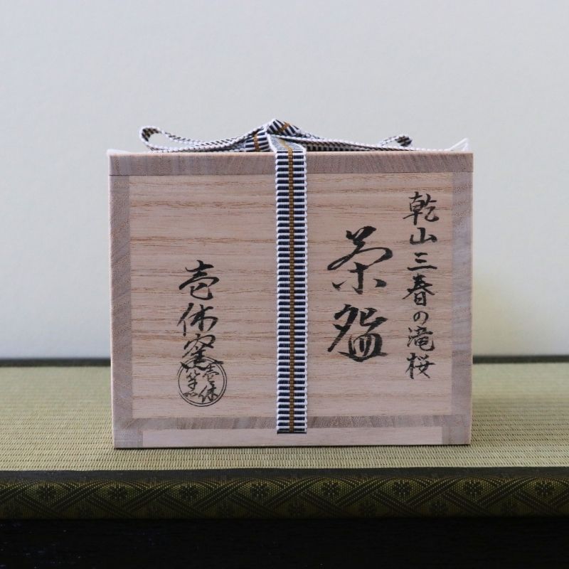 A light natural wood tea box with black Japanese calligraphy and a black-and-white striped ribbon tied around it, placed on a green textured surface against a plain light background.