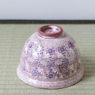A vintage Japanese ceramic bowl with a dusty pink glaze and purple floral patterns, photographed from underneath on a green woven mat.