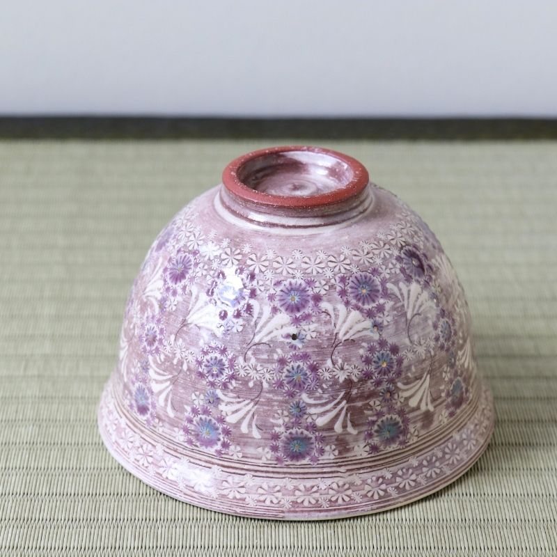 A vintage Japanese ceramic bowl with a dusty pink glaze and purple floral patterns, photographed from underneath on a green woven mat.